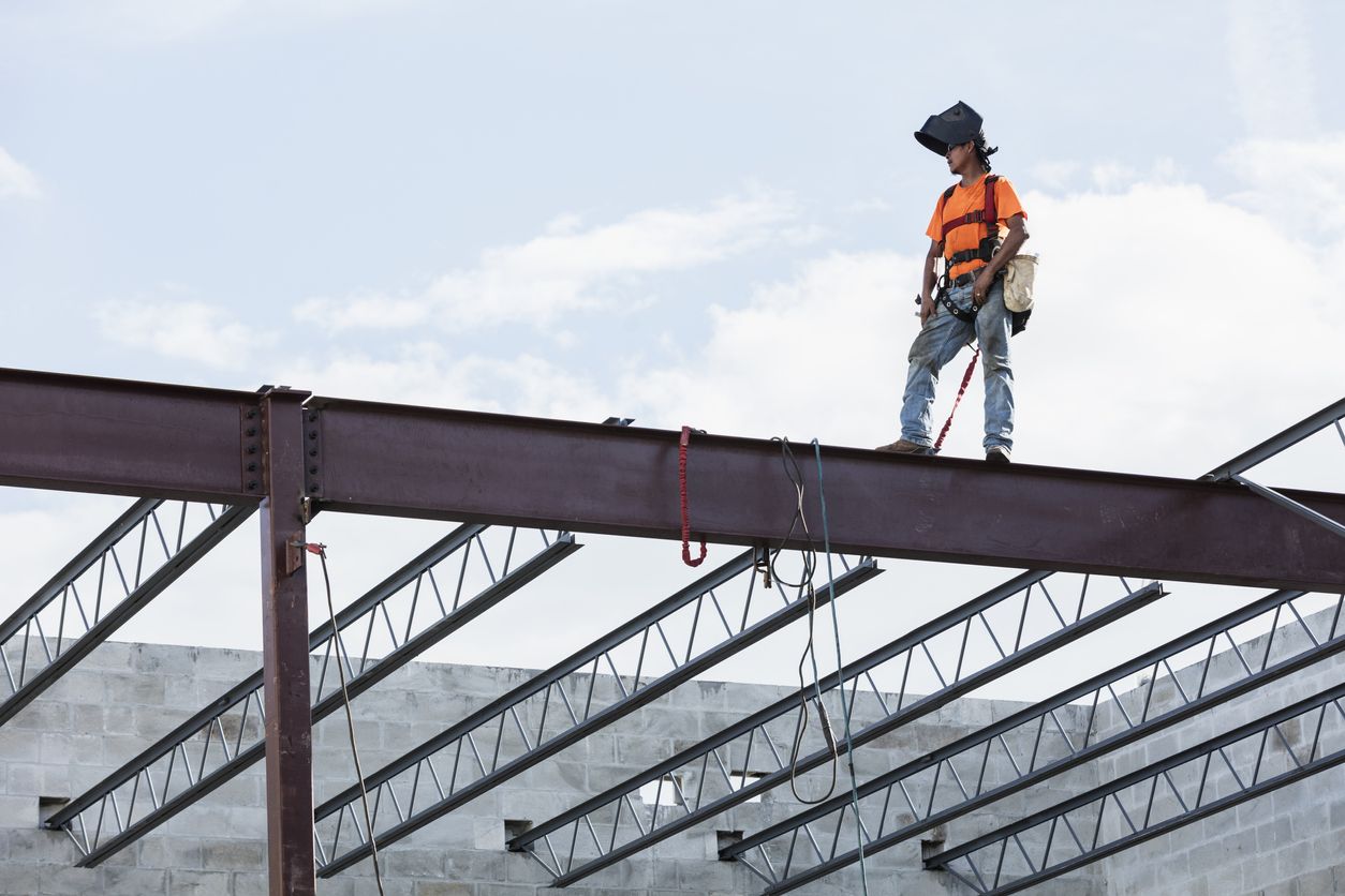 A Hispanic worker stands high up on a girder while wearing a flipped-up welding mask and safety harness.